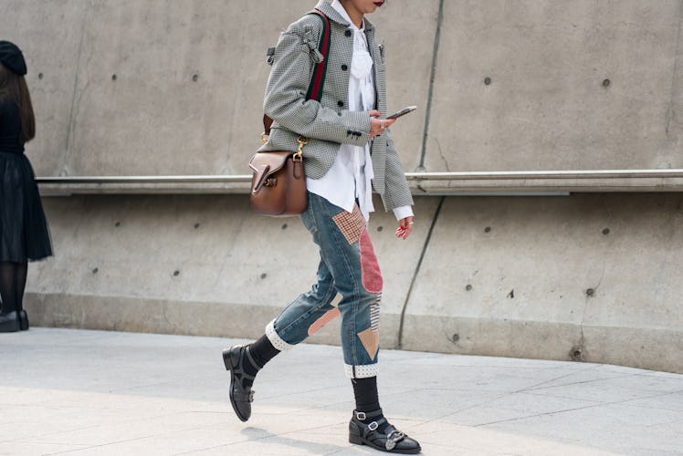 A girl in denim jeans with patches, a white shirt, and a grey jacket, looking at her phone while wal...