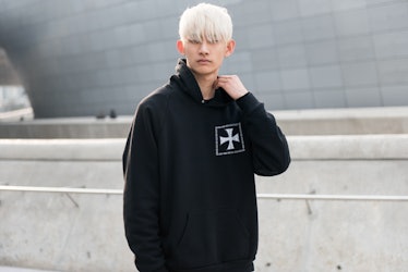 A blonde boy wearing a black hoodie with white cross posing for a photo during Seoul Fashion Week.