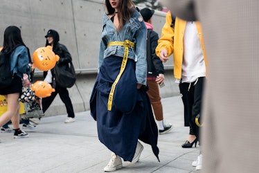 A woman in a blue dress, a denim jacket with a yellow tape measurer belt walking during Seoul Fashio...
