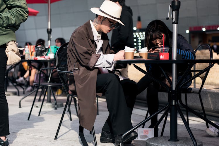 A man in a white shirt, a brown trench coat, and a white Fedora hat having a coffee during Seoul Fas...