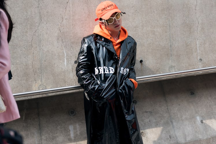 A woman wearing an orange cap, a matching hoodie with a faux black leather coat over during Seoul Fa...