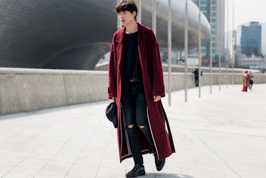 A man wearing a black t-shirt, black ripped skinny jeans and a maxi burgundy coat during Seoul Fashi...