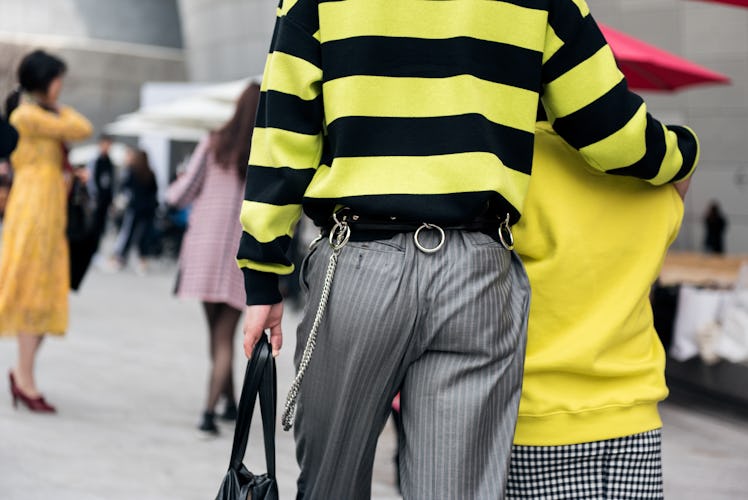 A man in a black-and-yellow sweater, grey slacks with chain details hugging a person in a yellow swe...