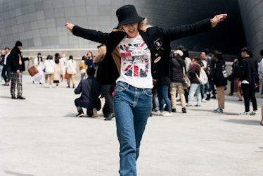 A woman wearing blue jeans, a white t-shirt with the UK flag, a black hat, and a blazer, joyfully sp...