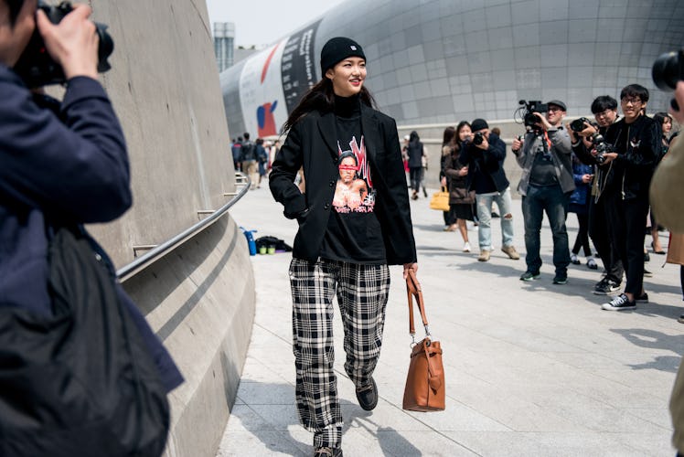 A woman wearing a black beanie, a graphic tee with plaid pants posing for a photographer in Seoul.