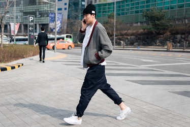 A man wearing a black beanie, a grey bomber jacket over a white shirt, black track pants crossing th...