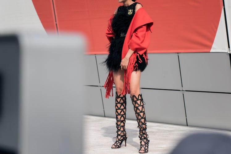 Stylish woman in black gladiator heels, fur dress, and red jacket, posing for a photo.