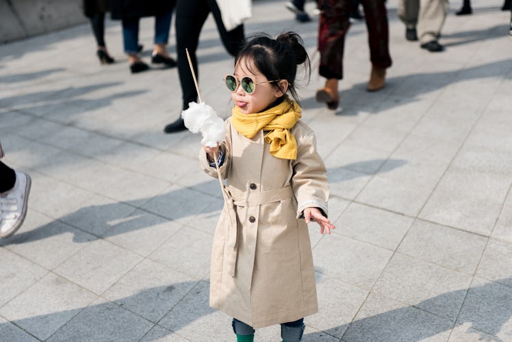 A toddler in a beige trench coat, yellow scarf and sunglasses holding a cotton candy on the street i...