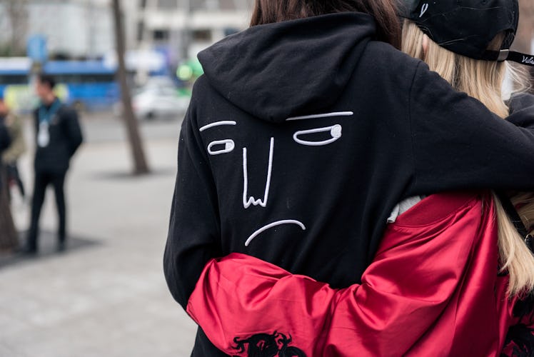 A woman in a black hoodie with face painted on its back hugging her friend.