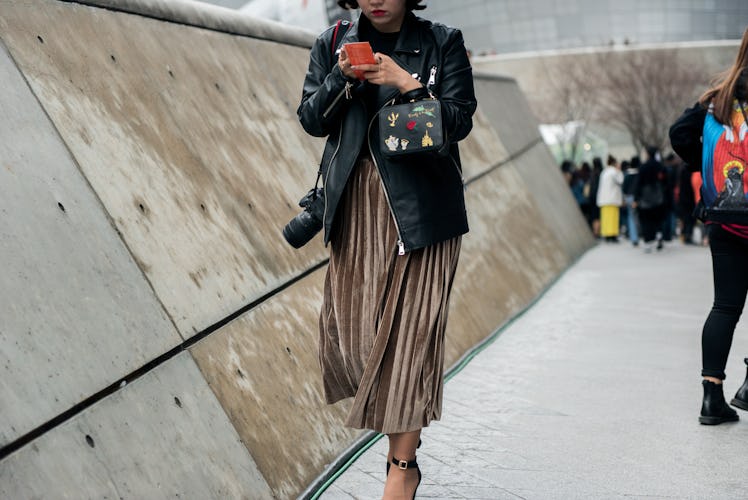 A woman in a beige pleated skirt, black leather jacket typing on her phone while walking the street ...