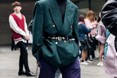 A man in blue pants, and an emerald-green blazer with a belt adorned with golden details walking the...
