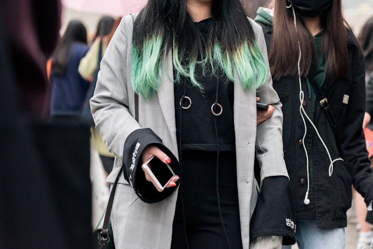 A woman with green highlights wearing a black hoodie with chain details and a grey coat