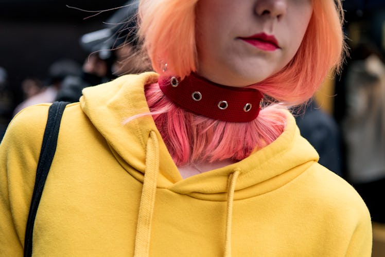 A woman with an orange and pink hair, wearing a yellow hoodie and a red choker necklace with punk de...