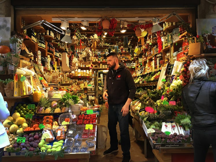 A man in a black shirt and trousers standing in the middle of a small grocery shop with various frui...