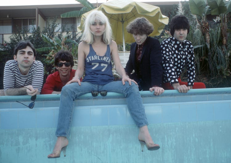 Debbie Harry sitting on a wall in a blue top and blue denim jeans with the members of the band Blond...
