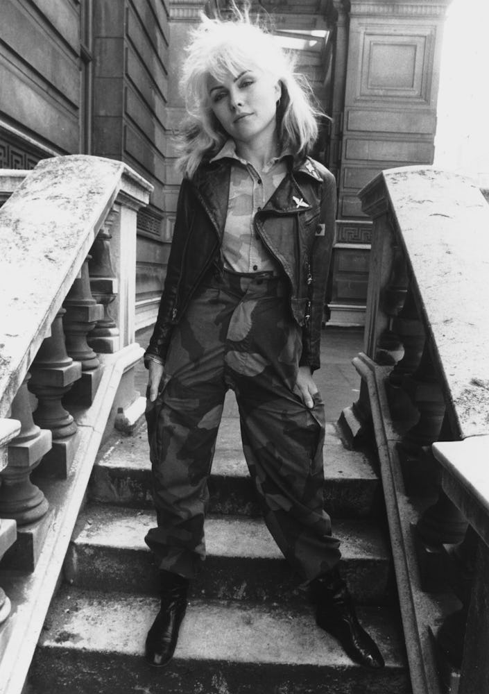 Debbie Harry standing in a shirt, leather jacket and camo print pants in black-and-white
