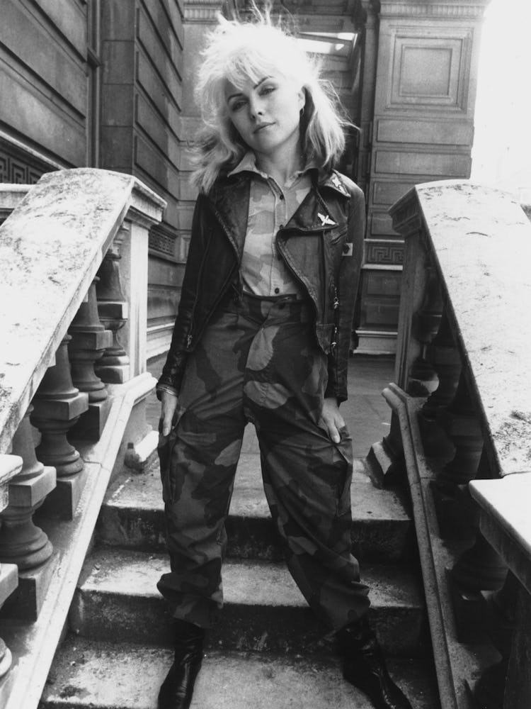 Debbie Harry standing in a shirt, leather jacket and camo print pants in black-and-white