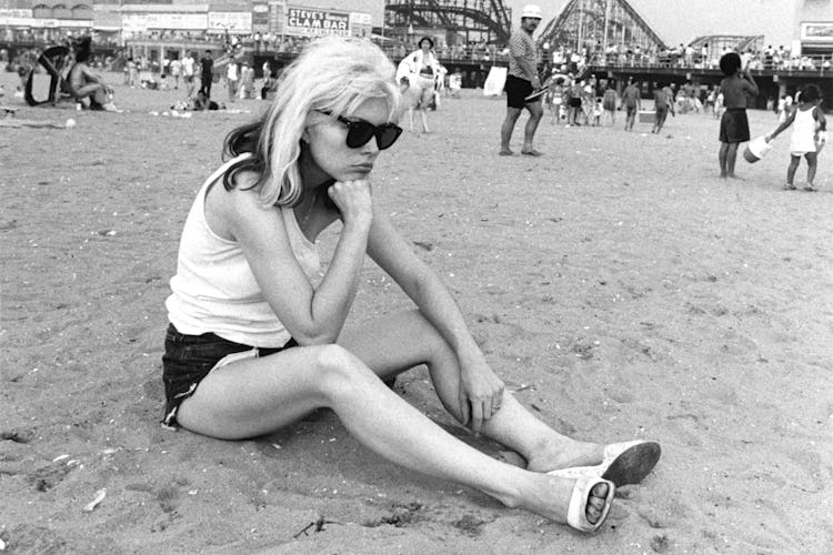 Debbie Harry in a white top, black shorts and sunglasses sitting on a beach and looking down