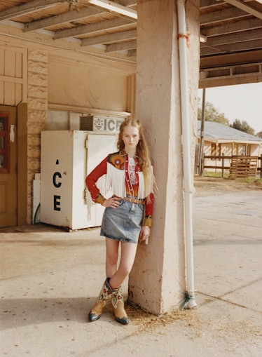 Jean Campbell leaning on a pillar while wearing a cowboy leather jacket, cowboy boots, and a jeans s...