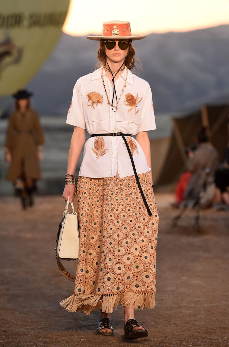 A female model walking while wearing a white shirt, brown dress, and a brown hat