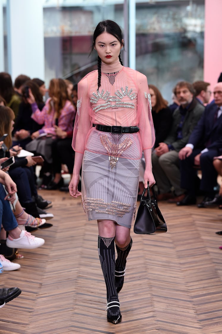 A black-haired female model walking a runway while wearing a pink blouse and a denim skirt