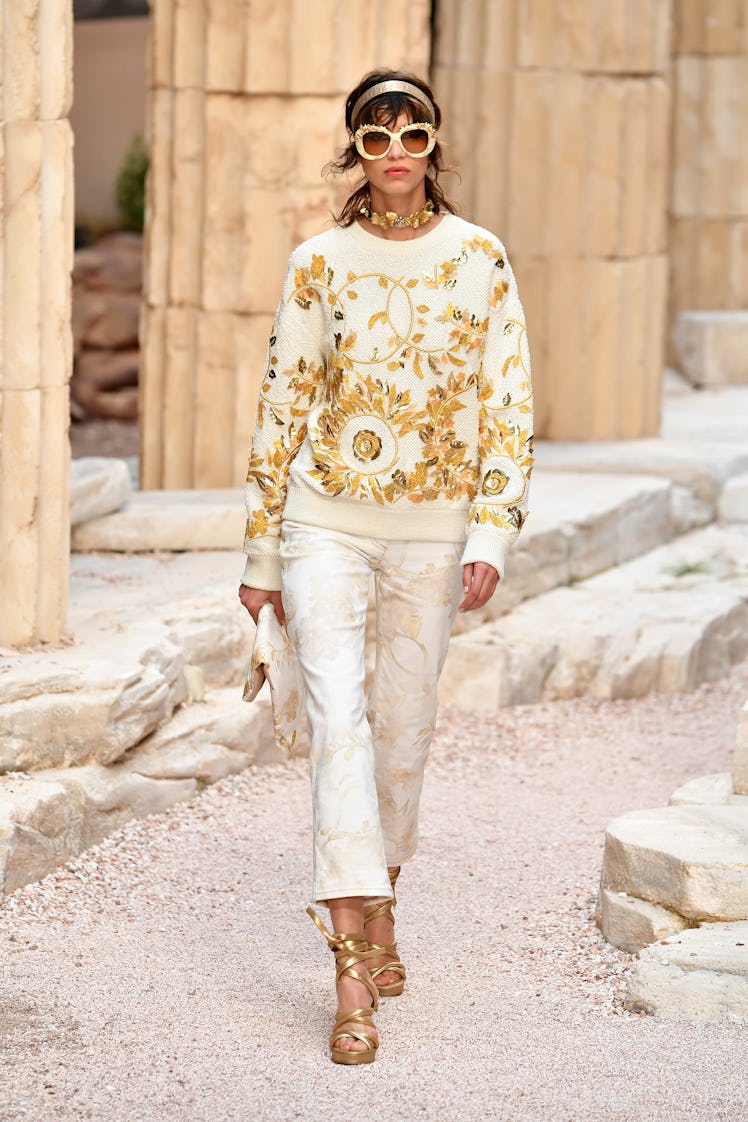 A female model walking while wearing oversized sunnies, a golden headband, and a white and golden ch...