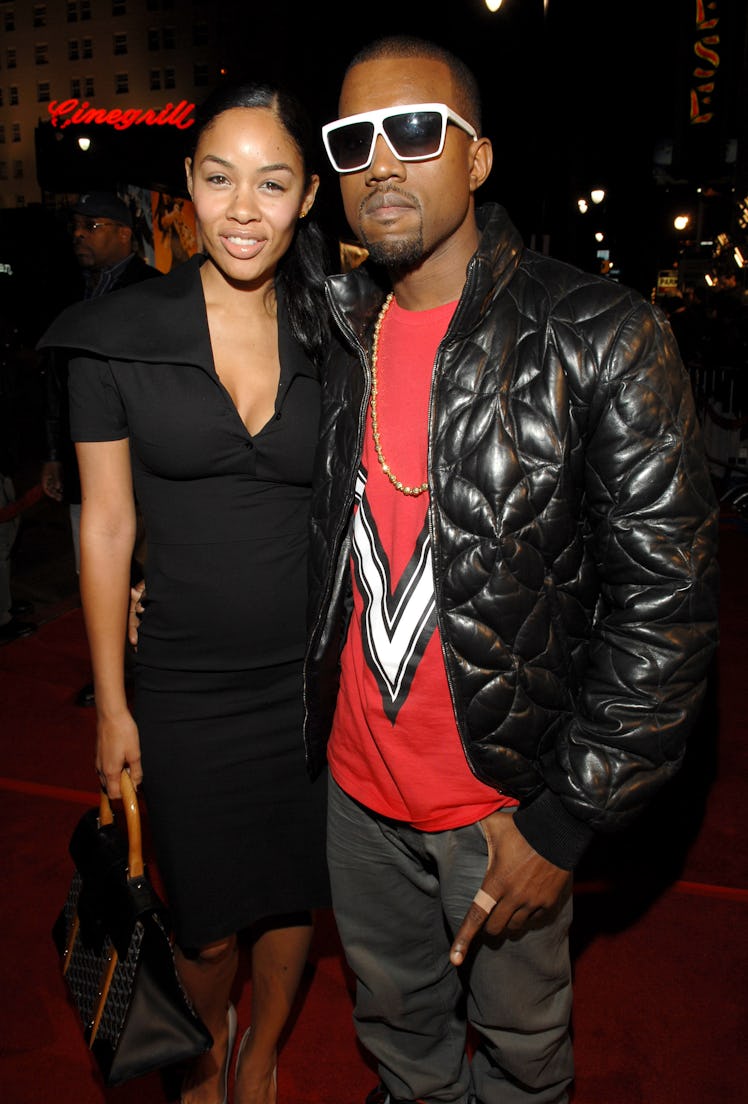 Kanye West in a red shirt, black jacket and grey trousers posing next to a woman at a red carpet eve...