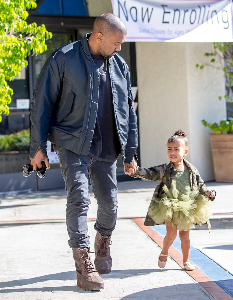 Kanye West in a black bomber jacket and shirt, grey pants and brown boots walking North West in a gr...