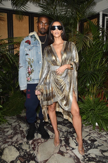 Kanye West in a floral denim jacket and navy pants and Kim Kardashian in a silver-gold dress and sun...