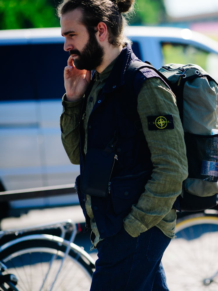A bearded man in a black-green jacket and trousers speaking on his phone and walking
