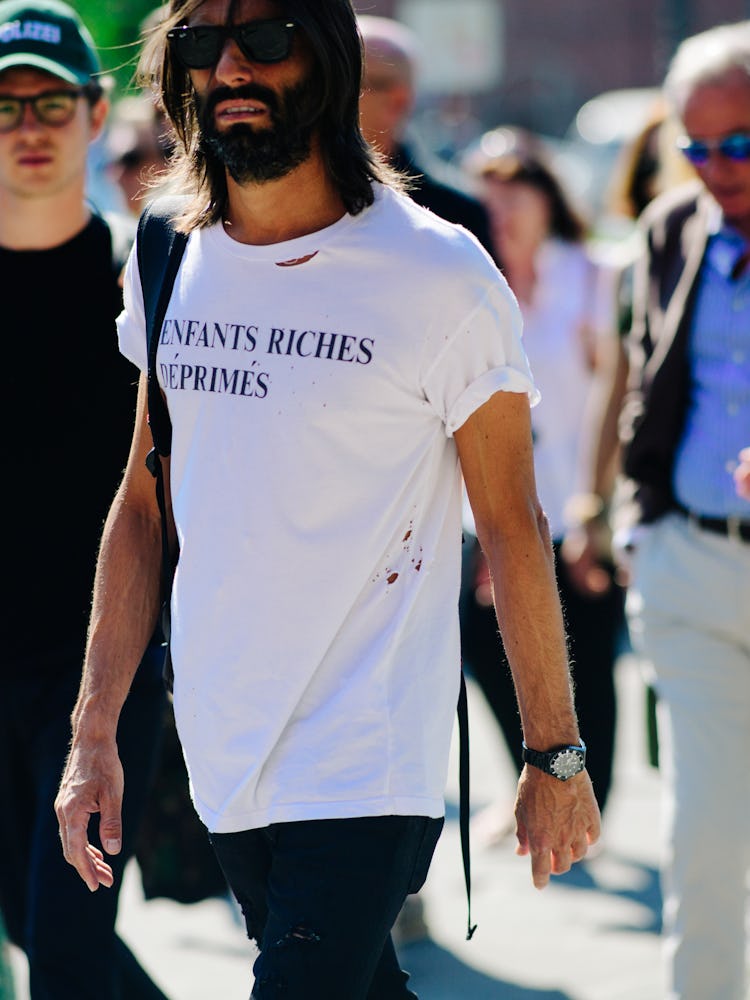 A bearded man in a white shirt with the print 'ENFANTS RICHES DEPRIMES' and black trousers