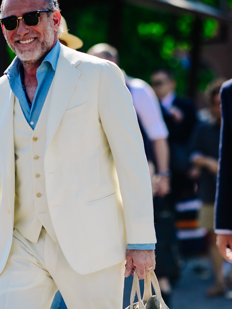A man walking in a white suit with waistcoat and a blue shirt whilst smiling