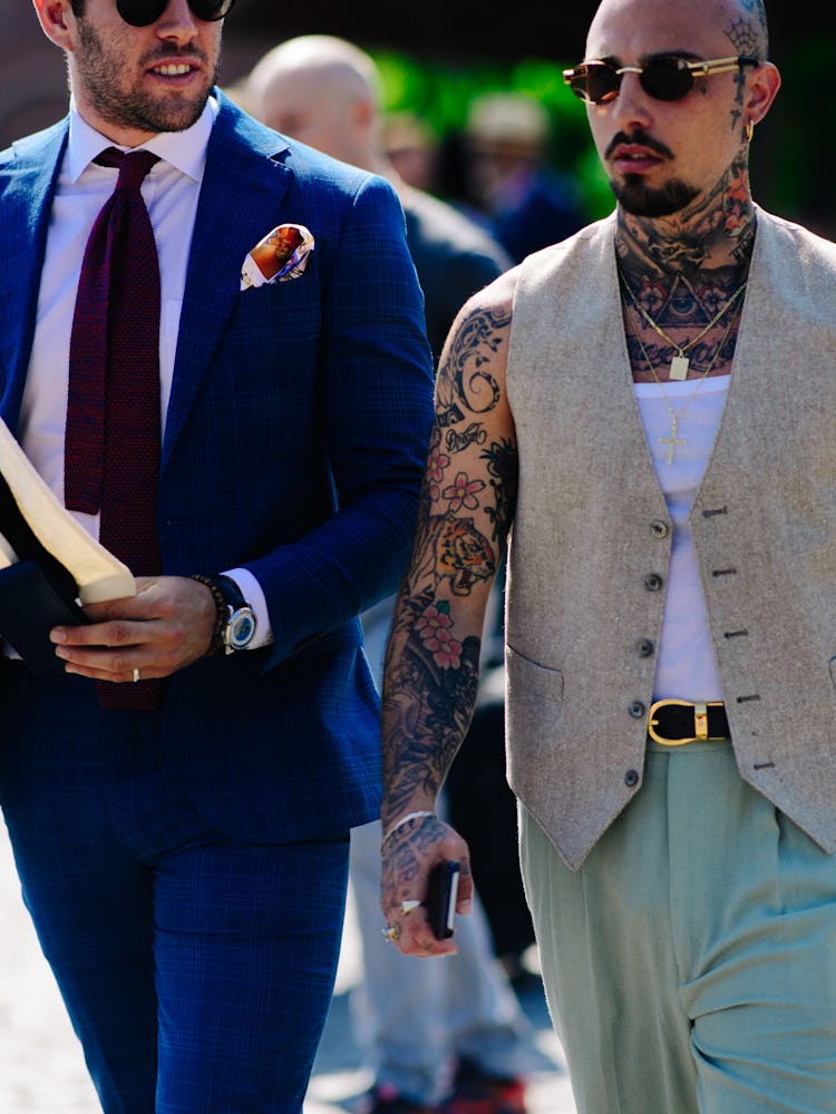 A man walking in a white top, a blazer waistcoat and sage trousers with tattooed arms and neck