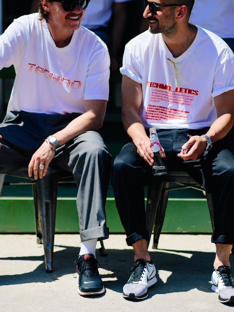 Two men in white shirts with red print and black pants sitting on a bench and having a conversation