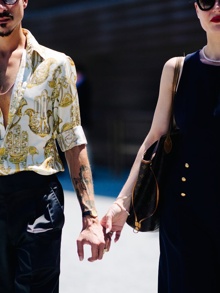 A man in a white-gold shirt and black pants and a woman in a black dress holding hands and walking