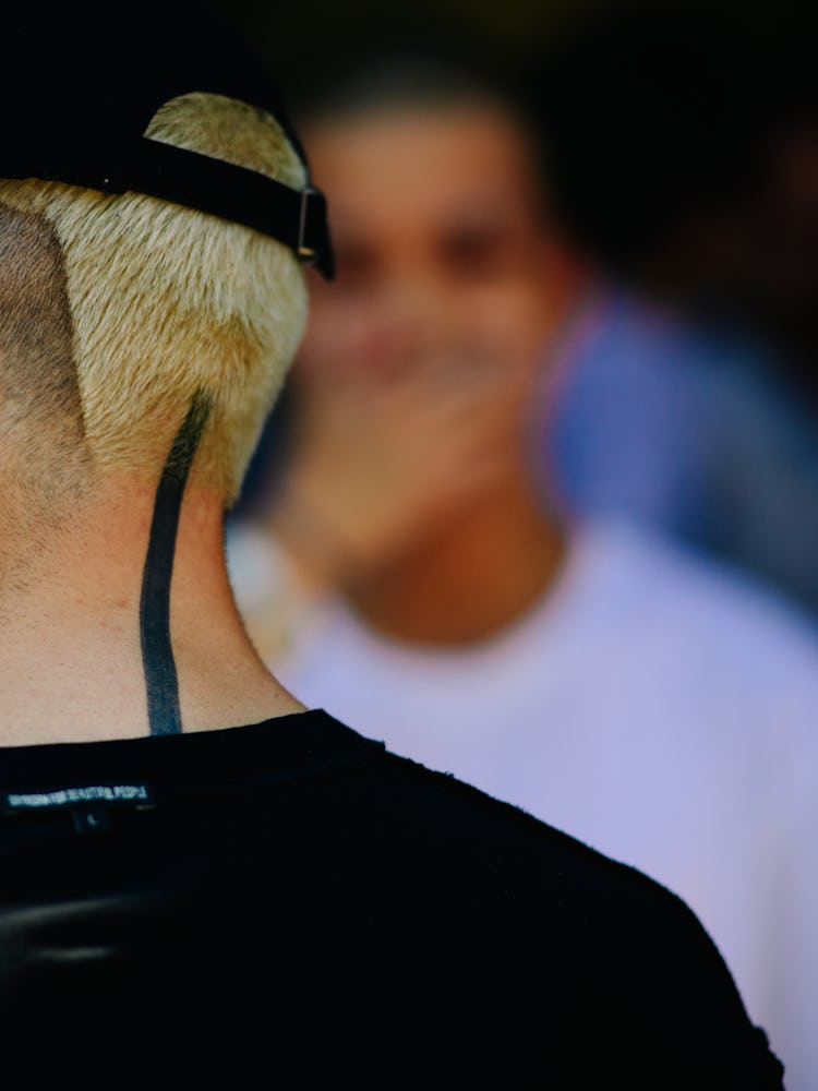 The back of a blonde man in a black shirt and a black cap with a tattoo on the back of his neck