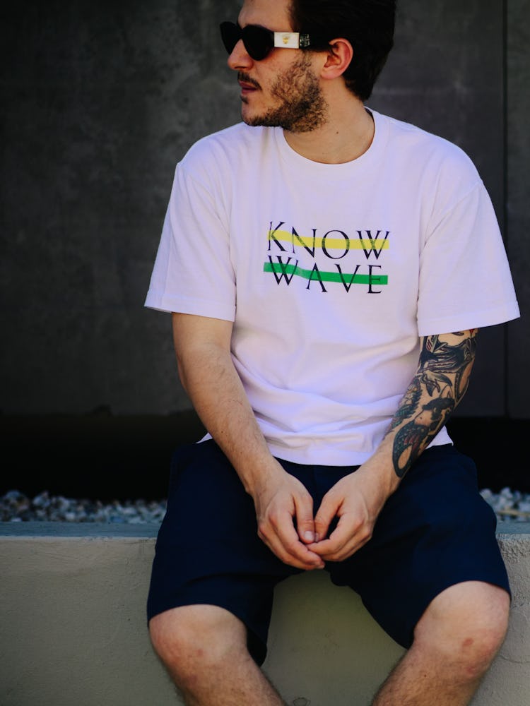 A man in a white shirt with the printed text 'KNOW WAVE', black shirt and sunglasses sitting on a be...