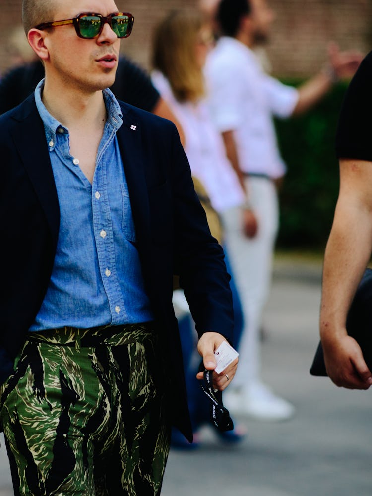 A man in a blue denim shirt, black blazer and green-black trousers with leaf print