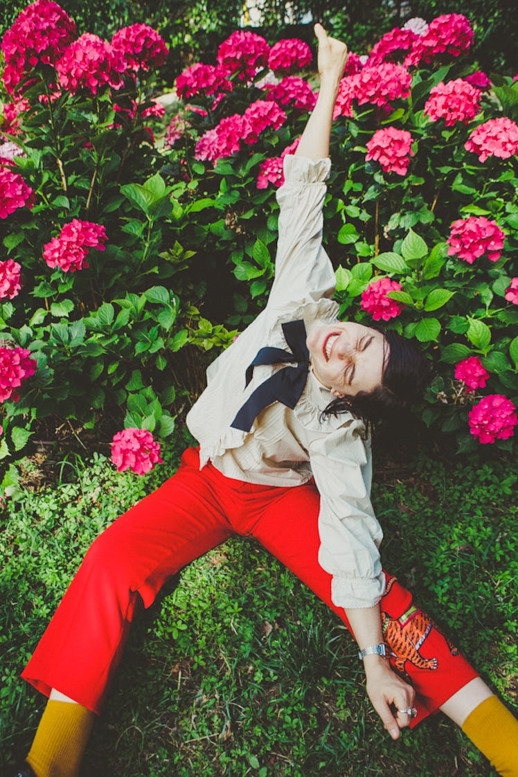Soko wearing red pants and a white shirt with a black bow tie posing next to a flower bush.