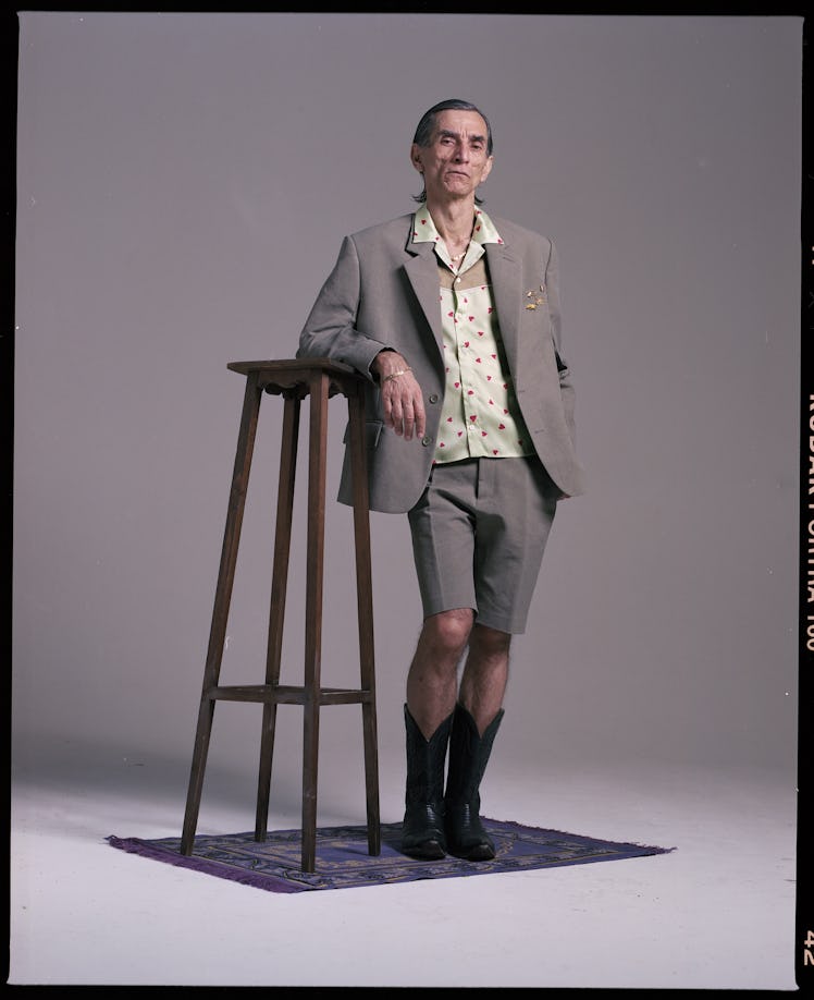 A man wearing a grey blazer and shirt and a beige shirt by Magliano leaning against a barstool