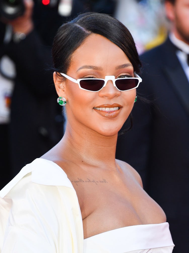 Rihanna wearing a slicked back up do with her a side part, white sunglasses, minimalistic makeup and...