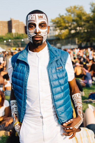 A man with white face paint, in a white shirt and pants. and a blue puffer vest at the Afropunk Fest...