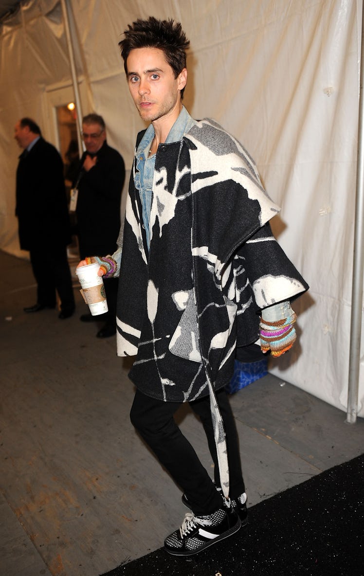 Jared Leto in a black-white-grey poncho and black trousers walking while holding a cup of coffee