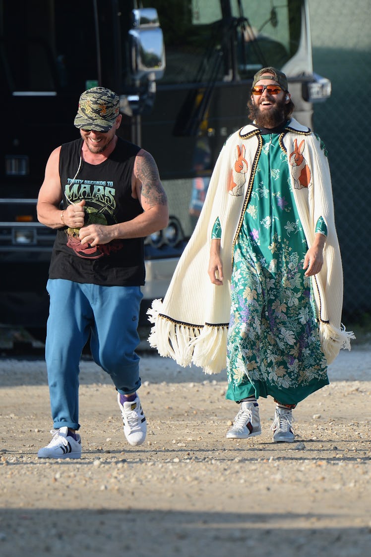 Jared Leto in a green floral dress and a beige floral poncho walking next to a man in a black top an...