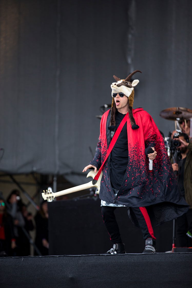 Jared Leto in a black-red poncho and pants and a goat plush headpiece holding a guitar on stage