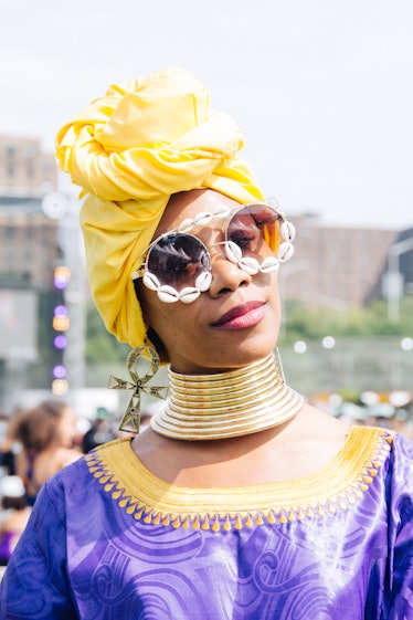 A woman wearing a yellow headpiece, sunglasses with shells on them, a gold statement necklace and pu...