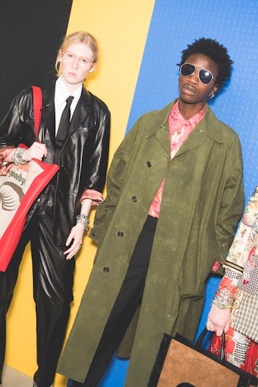 A model in a green trench coat and a model in a leather suit and black tie posing backstage at the G...