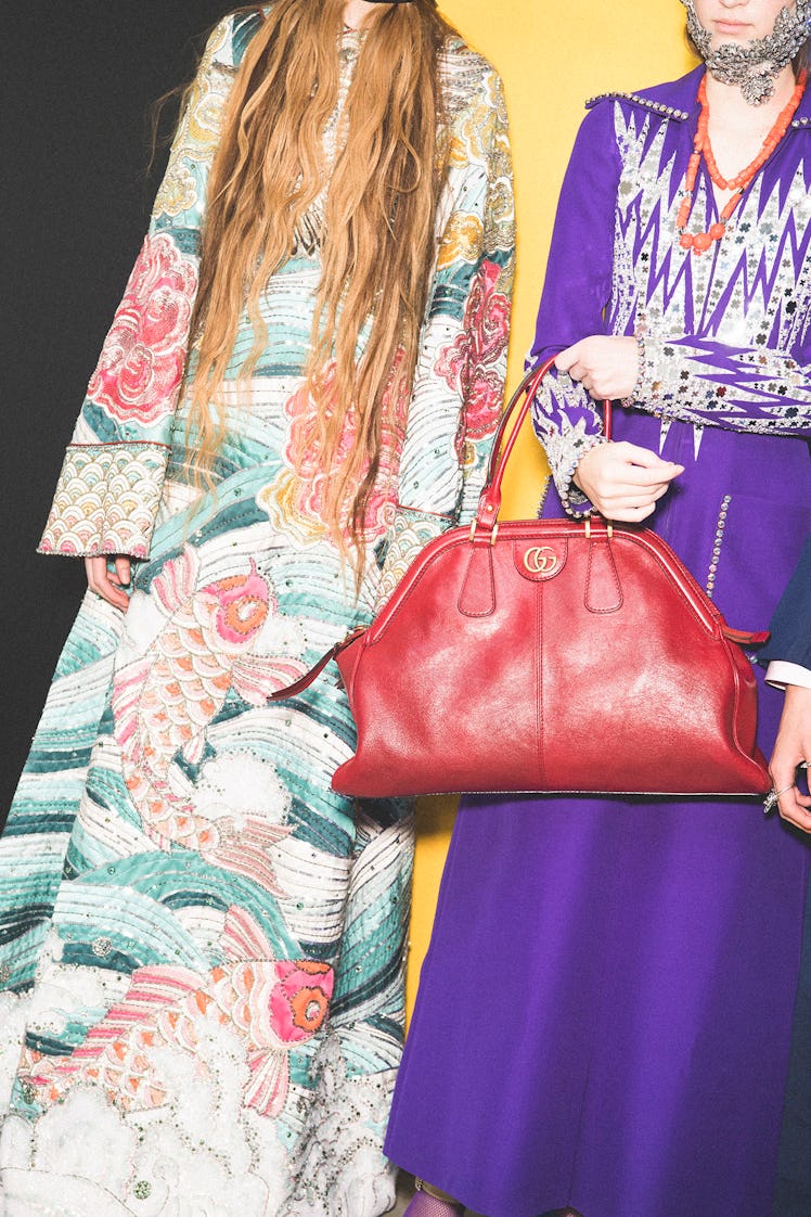 Two models backstage at the Gucci show in floor-length gowns, one's is purple and the other's has na...