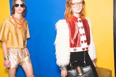 Two models walking backstage at the Gucci show one in a shimmery top and the other in a red leather ...