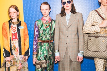 Models posing backstage at the Gucci show in floral gowns and one in a beige suit with a pleated ski...